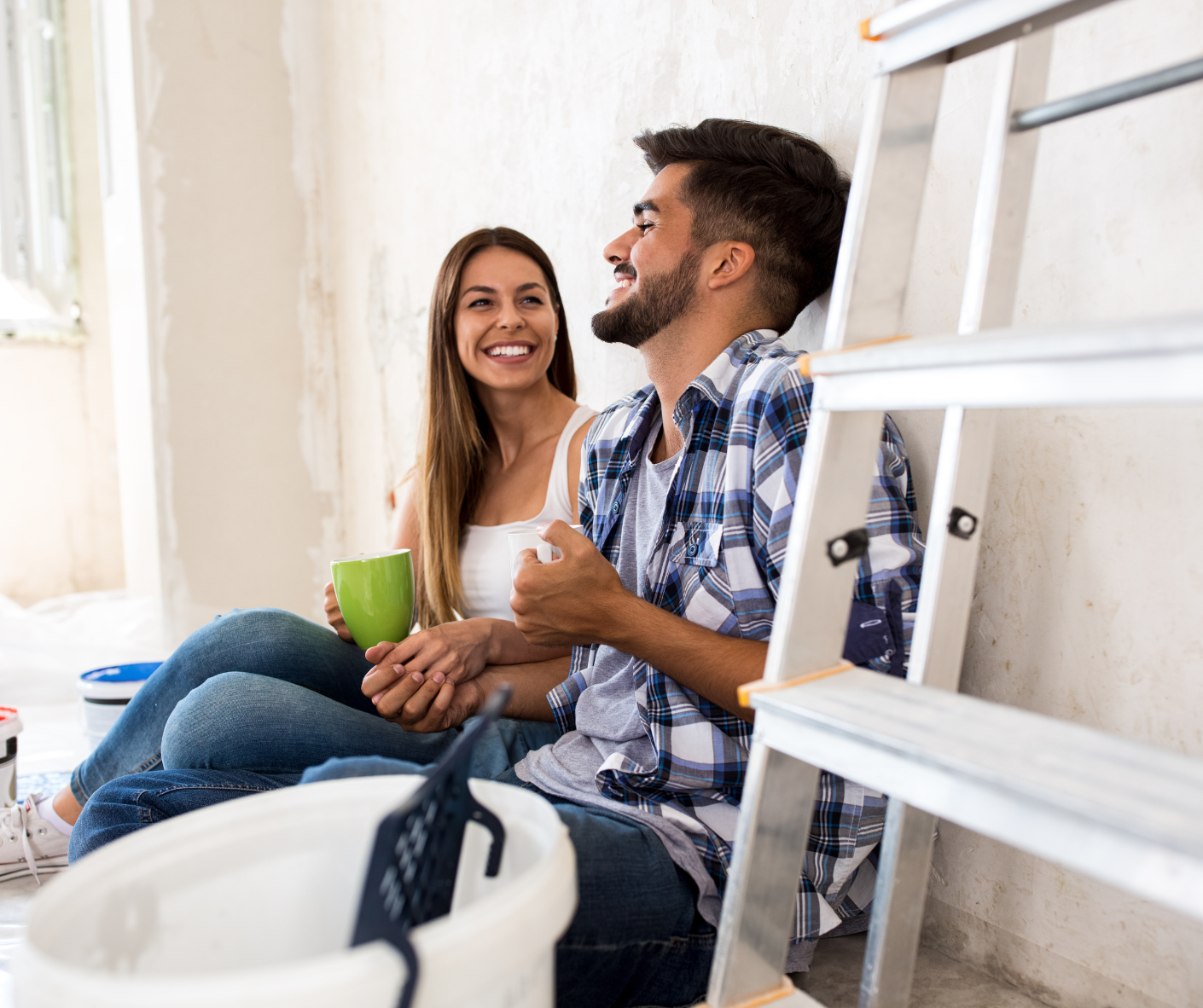 Couple assis au sol dans une maison en renovations, avec tasse de café à la main, souriants